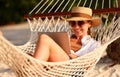 Young smiling female, happy woman in straw hat and sunglasses using digital tablet while relaxing in the hammock on tropical sandy Royalty Free Stock Photo