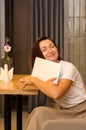 Young, happy, tired caucasian woman with brown hair sits in light cafe at wooden table with flower, hugging white laptop Royalty Free Stock Photo