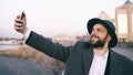 Young happy tourist man in ahat and coat smiling while taking selfie picture with mobile phone on city riverside Royalty Free Stock Photo