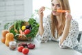 Young and happy woman with carrot at table with green fresh ingredients indoors.