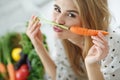 Young and happy woman with carrot at table with green fresh ingredients indoors.