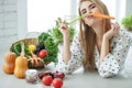 Young and happy woman with carrot at table with green fresh ingredients indoors.