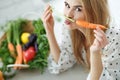 Young and happy woman with carrot at table with green fresh ingredients indoors.