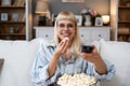 Young happy woman changing channels with remote control while watching TV and eating popcorn in the evening at home. Relaxed