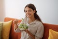 Young happy woman eating vegetable salad while sitting on couch at home Royalty Free Stock Photo