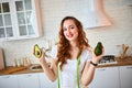 Young happy woman holding avocado for making salad in the beautiful kitchen with green fresh ingredients indoors. Healthy food and Royalty Free Stock Photo