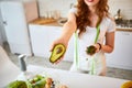 Young happy woman holding avocado for making salad in the beautiful kitchen with green fresh ingredients indoors. Healthy food and Royalty Free Stock Photo