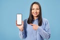 A young, happy woman is holding a smartphone and pointing at a blank white screen on a blue background Royalty Free Stock Photo