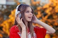 Young happy woman listening to music on headphones standing outdoors on autumn colorful leaves background