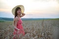 Young happy woman in red summer dress and white straw hat walking on yellow farm field with ripe golden wheat enjoying warm Royalty Free Stock Photo
