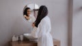 Young happy woman standing in bathroom looking in mirror touching face after applying cream enjoys healthy moisturized Royalty Free Stock Photo