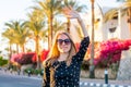 Young happy woman in sunglasses waves her hand in front of blooming red and pink hibiscus and palm trees at sunset Royalty Free Stock Photo