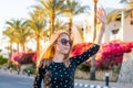 Young happy woman in sunglasses waves her hand in front of blooming red and pink hibiscus and palm trees at sunset Royalty Free Stock Photo