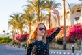 Young happy woman in sunglasses waves her hand in front of blooming red and pink hibiscus and palm trees at sunset Royalty Free Stock Photo