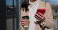 Young happy woman using smartphone and drinking coffee to go outdoors, walking down the city street. Royalty Free Stock Photo