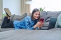 Young happy woman using smartphone laptop, lying on sofa at home Royalty Free Stock Photo