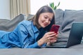 Young happy woman using smartphone laptop, lying on sofa at home Royalty Free Stock Photo