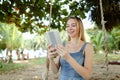 Young happy woman using tablet and riding swing on sand, wearing jeans sundress. Royalty Free Stock Photo