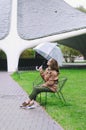Young happy woman walking in the rain in green park with umbrella, smiling, fun. person sitting on the bench. Cold weather Royalty Free Stock Photo