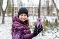 Young happy woman in warm purple dawn jacket smiling while making selfie with smartphone during walk in winter city park