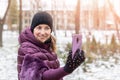 Young happy woman in warm purple dawn jacket smiling while making selfie with smartphone during walk in winter city park. Pretty