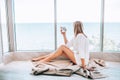 Young happy woman in a white knitted dress enjoying sea view next to big window with cup of tea. Panoramic window. Floor covering Royalty Free Stock Photo