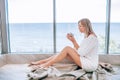 Young happy woman in a white knitted dress enjoying sea view next to big window with cup of tea. Panoramic window. Floor coverings Royalty Free Stock Photo