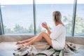 Young happy woman in a white knitted dress enjoying sea view next to big window with cup of tea. Panoramic window. Floor coverings Royalty Free Stock Photo