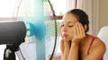 Young headache woman suffering a heat wave and using a fan sitting on a couch in the living room at home