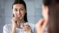 Young healthy woman holding pill water glass looking in mirror