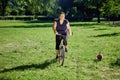 Young healthy woman rides bike in park with small dog running near. Royalty Free Stock Photo