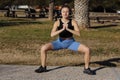 Young healthy woman in sportswear listens music in headphones while doing fitness squat leg exercises outdoors in park Royalty Free Stock Photo