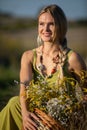 A young herbalist crouches by the dirt road with a basket full of herbs and looks into the sun.