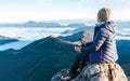 Young hiker female sitting on the mountain summit cliff and enjoying mountains valley covered with clouds view. Successful summit