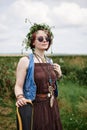 Young hippie woman with short red hair, wearing boho style clothes, sunglasses and flower wreath, standing on green field, holding Royalty Free Stock Photo