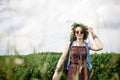 Young hippie woman with short red hair, wearing boho style clothes, sunglasses and flower wreath, standing on green field, holding Royalty Free Stock Photo