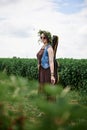 Young hippie woman with short red hair, wearing boho style clothes, sunglasses and flower wreath, standing on green field, holding Royalty Free Stock Photo