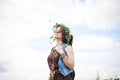 Young hippie woman with short red hair, wearing boho style clothes, sunglasses and flower wreath, standing on green field, holding Royalty Free Stock Photo