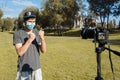 Young hipster blogger, wearing a protective medical mask, recording a video blog on a digital camera in the park. Lead blogger.