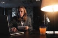 Young hipster businessman sits by the wooden table with lamp, laptop and smartphone at night office. Man with long hair work with