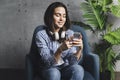 Young hipster girl sitting in armchair and texting on smartphone. Entertaining and educating content online. Female communicating