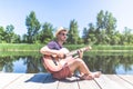 Young hipster guy holding acoustic guitar while sitting on dock in nature. Summer and travel concept Royalty Free Stock Photo