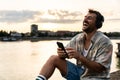 Young hipster happy man sitting near the river in the city escaping from city life listening music headset enjoying peace and good