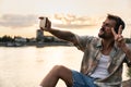 Young hipster happy man sitting near the river in the city escaping from city life listening music headset enjoying peace and good