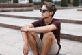 Young hipster man in black sunglasses with a stylish hairstyle in a brown t-shirt in summer shorts sits on the vintage steps Royalty Free Stock Photo