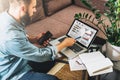 Young hipster man, entrepreneur sits at home on couch at coffee table, holding smartphone, showing pencil on screen