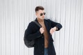 Young hipster man in stylish sunglasses in a classic black coat in jeans in a knitted vintage sweater with a backpack outdoors Royalty Free Stock Photo