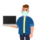 Young hipster man wearing medical mask and showing laptop computer. Trendy person covering face protection from virus. Royalty Free Stock Photo