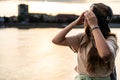 Young hipster millennial woman sitting on riverside looking at city listening music on wireless headphones enjoying moment, life Royalty Free Stock Photo