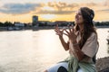 Young hipster millennial woman sitting on riverside looking at city listening music on wireless headphones enjoying moment, life Royalty Free Stock Photo